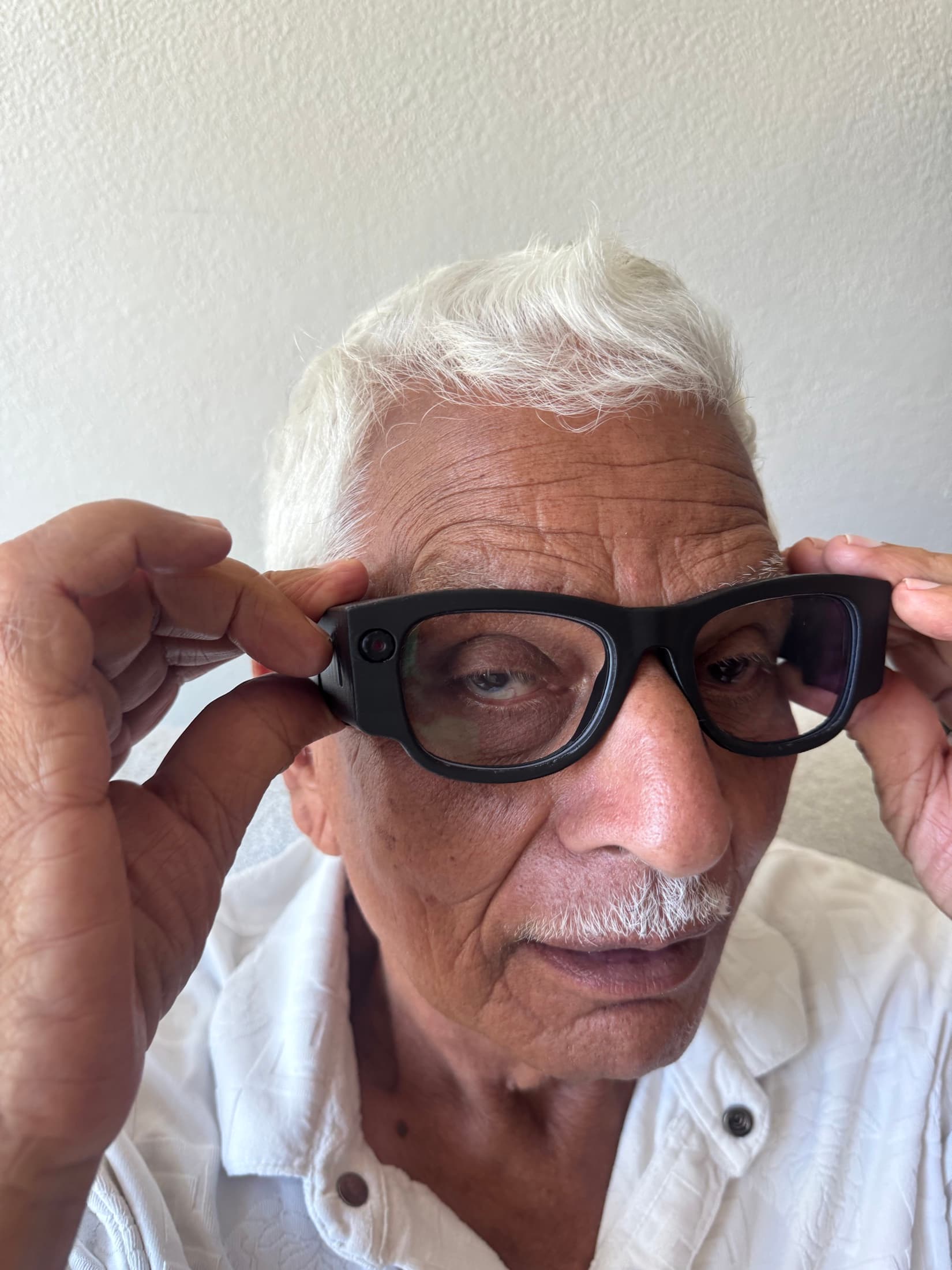 A man with white hair holds the EchoPath glasses with both hands as he puts them on, looking straight at the camera.